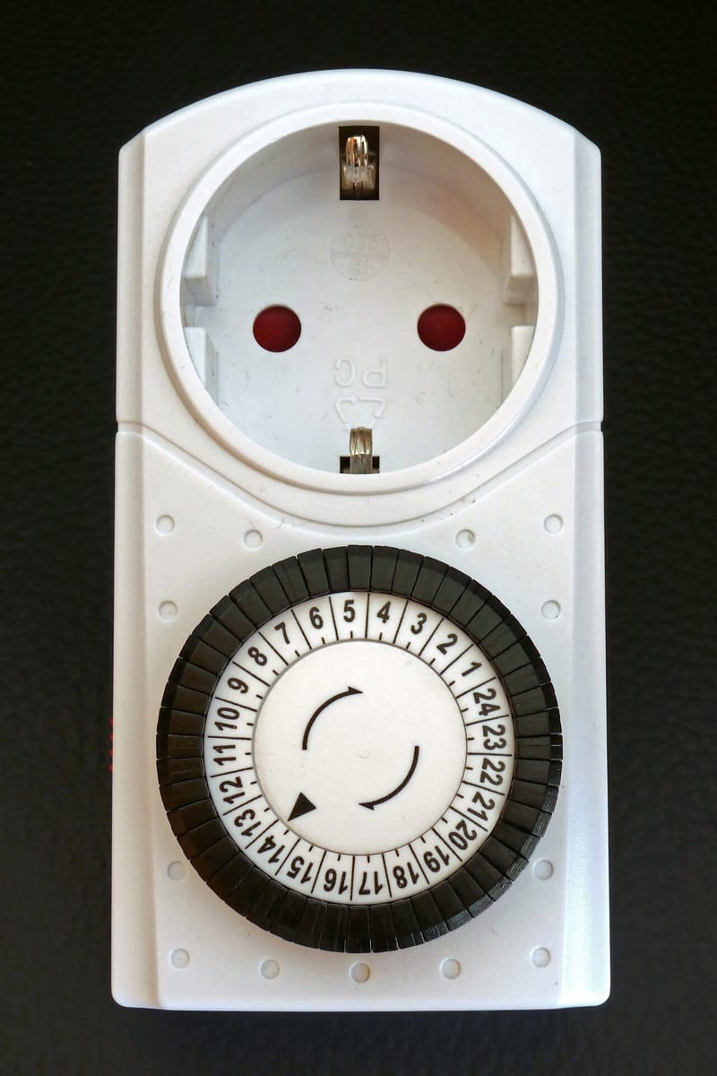 A white mechanical plug-in outlet timer with a 24-hour dial and segmented pins around the rim.