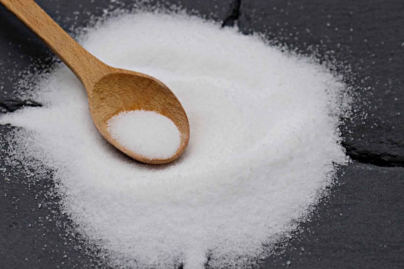 A wooden spoon holding coarse white salt crystals against a dark surface, illustrating a tablespoon-scale dose.