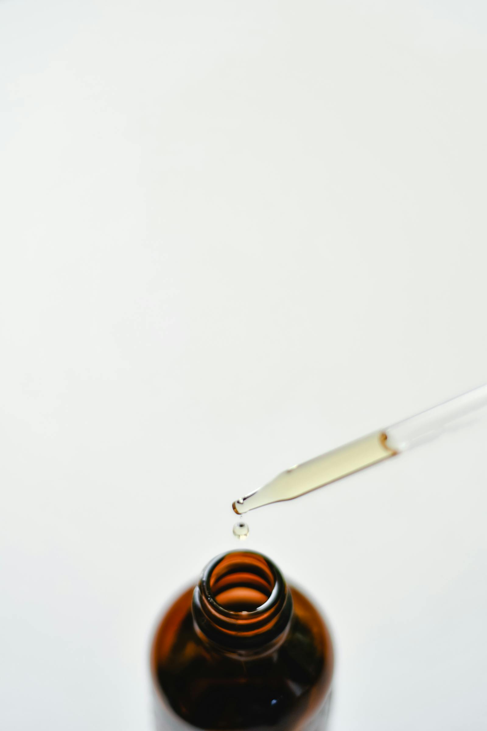 Brown glass dropper bottle with a single droplet of amber liquid hanging from the glass pipette, on a neutral white background.